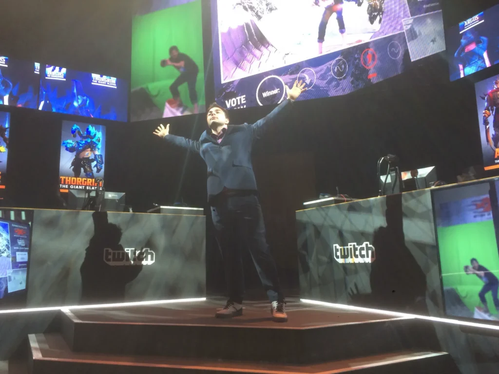 Ein Announcer steht mit ausgebreiteten Armen auf einer Messebühne mit Twitch-Logos und großen Bildschirmen