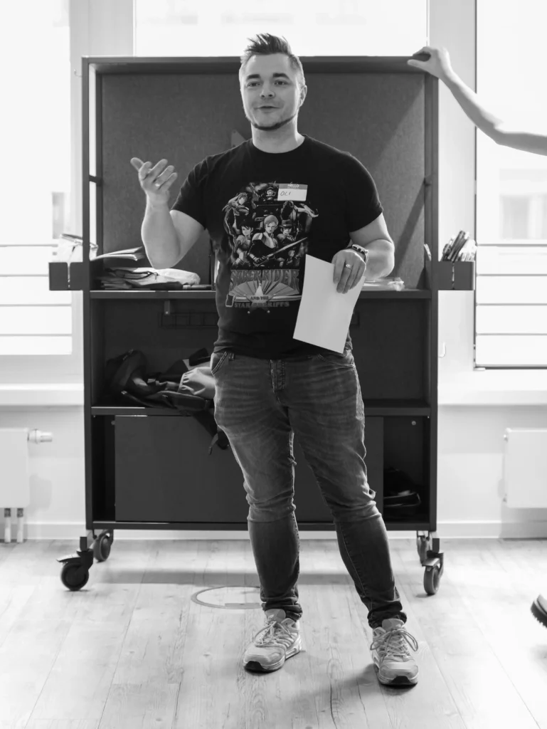 Oli spricht mit Namensschild auf dem T-Shirt und einem Blatt Papier in der Hand vor einer Gruppe
